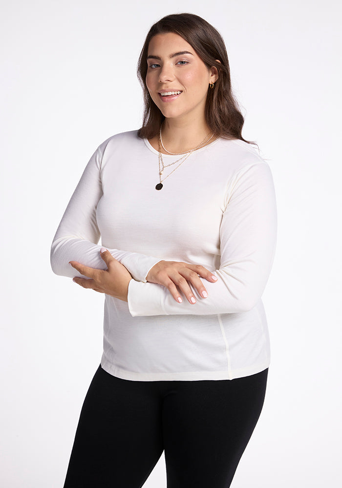 A woman with long brown hair wearing a Woolx Hannah Long Sleeve Top in buttercream and black pants. She accessorizes with gold necklaces and poses with her arms crossed against a white background. | Juliana is 5'10", wearing a size XL